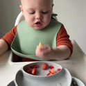 Baby in a high chair with a green bib and a bowl of fruits.